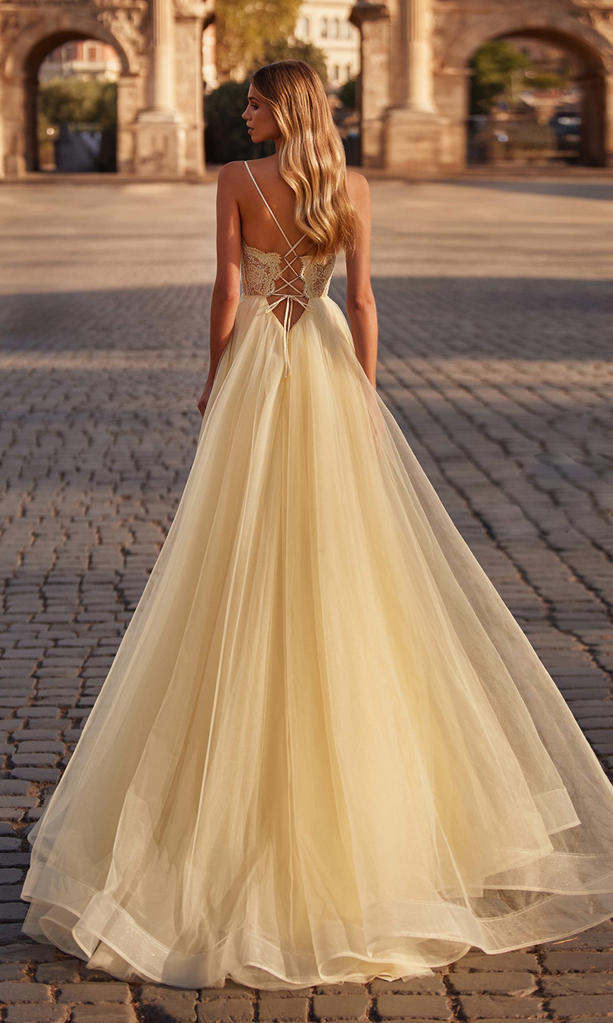 A woman in a flowing pale yellow La Femme Lace-Bodice Long A-Line Prom Dress 32306 with a lace-up back stands on a cobblestone street, facing away from the camera.